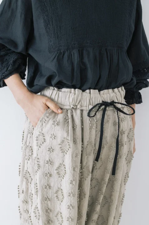 黒いピンタックブラウスとベージュの刺繍入りドローストリングパンツを合わせた、ナチュラルでリラックス感のある女性のコーディネート