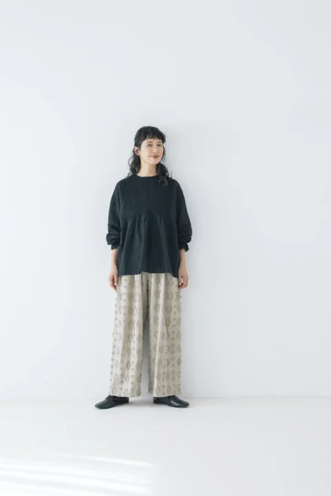 プリーツデザインの黒いトップスに、ベージュの柄入りワイドパンツと黒いフラットシューズを合わせた、ナチュラルでリラックス感のあるコーディネート