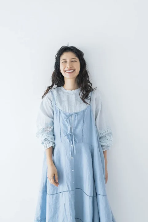 水色のワンピースとブラウスを重ね着した女性が、目を閉じて満面の笑みを浮かべているポートレート