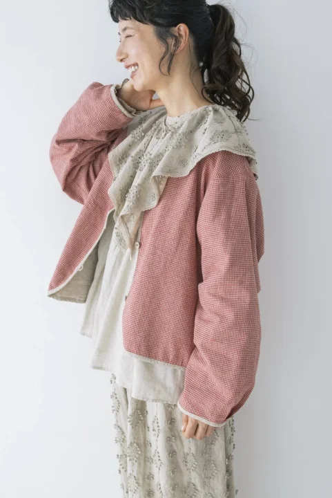 笑顔の女性が、赤と白のギンガムチェックのアウターと、フリルと刺繍が特徴的なブラウス、刺繍入りのベージュのスカートを着用し、ナチュラルな雰囲気のコーディネートを見せている