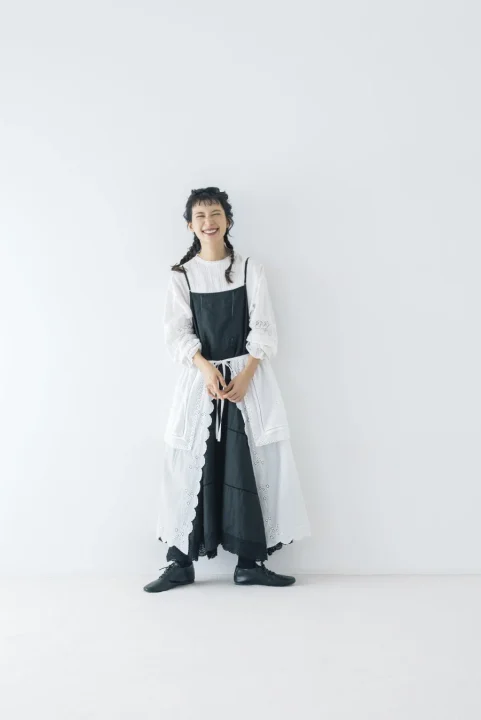 白いブラウスに黒いキャミソールワンピース、白いレースのベストを重ね着したナチュラルなファッションの女性