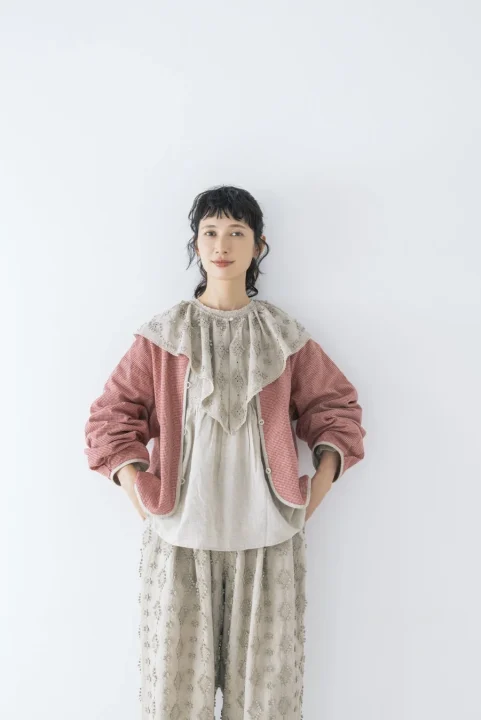 赤と白のギンガムチェック柄のジャケットを羽織り、ベージュの刺繍入りブラウスとパンツを着用したナチュラルな女性