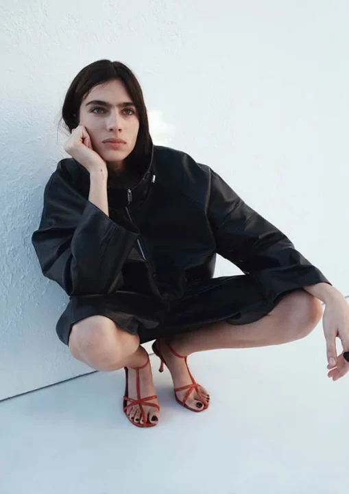 黒いレザージャケットと赤いストラップサンダルを身につけた女性が、白い背景の前でクールな表情でしゃがみ込むポーズ