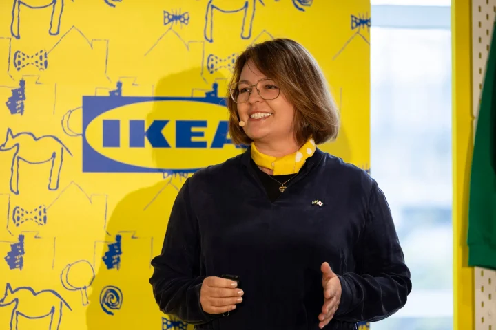 眼鏡をかけた女性が、イケアのロゴとイラストが描かれた黄色の壁を背景に笑顔で話している