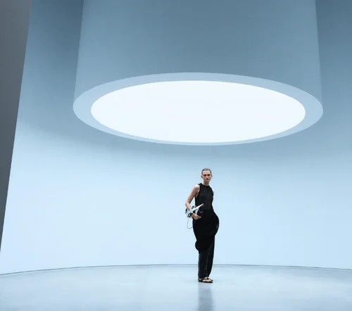 青い壁と円形の照明が特徴的なミニマルな空間に、黒い服を着たモデルが立っている。現代的で洗練された雰囲気の画像。