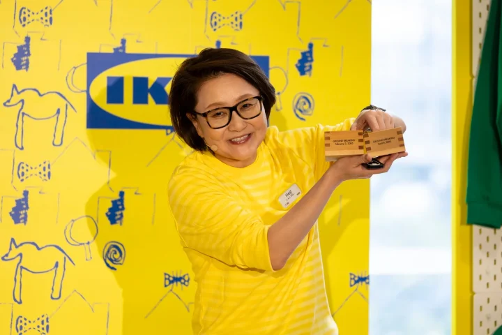 イケアのロゴを背景に、黄色いシャツを着た女性が笑顔で2つの木製記念品を持っている