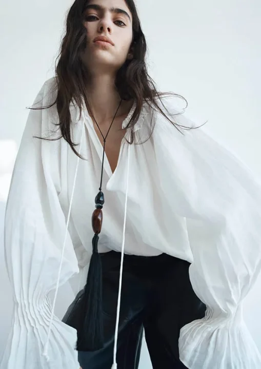 白いルーズフィットのブラウスと黒いパンツを着用し、長いタッセル付きのネックレスをアクセントにしたモデルのポートレート