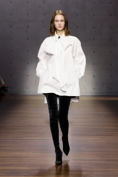 大きなリボンが特徴的な白いオーバーサイズシャツと、黒いサイハイブーツを着用したモデル