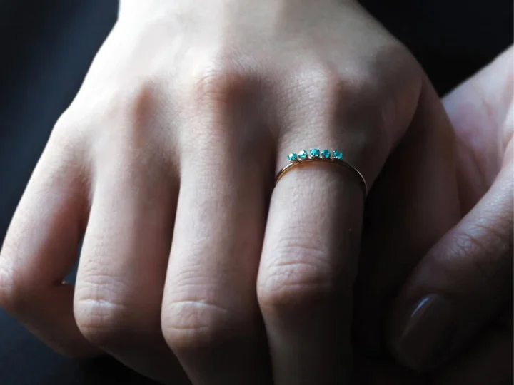 華奢な指に、複数の小さな青緑色の宝石があしらわれたゴールドの指輪をはめている様子を捉えた画像です。