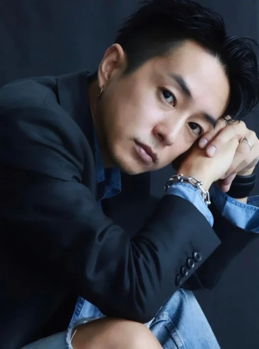 黒いジャケットとデニムシャツを着用し、カメラを見つめる男性のクローズアップポートレート。腕を組み、指輪やブレスレットなどのアクセサリーを身につけている。