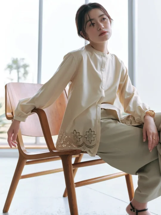 窓から自然光が差し込む明るい室内で、木製の椅子に座る若い女性のポートレート。ベージュのレース刺繍入りブラウスとオリーブグリーンのワイドパンツを着用し、落ち着いた表情で遠くを見つめています。