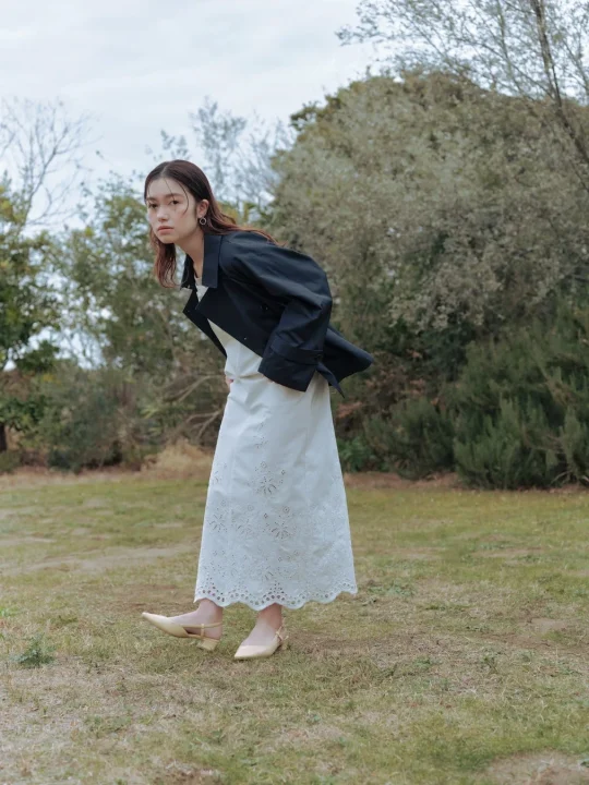 自然の中で、白い刺繍のロングワンピースに黒いジャケットを羽織った若い女性がカメラを見つめているポートレート写真です。