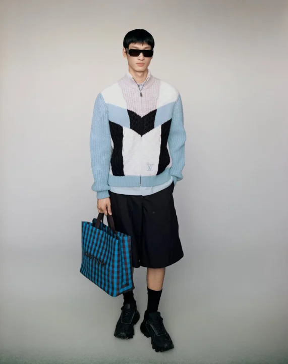 カラフルなルイヴィトンのセーターと黒いショーツを着用した男性モデルが、サングラスとスニーカーを合わせ、青と茶色のチェック柄のルイヴィトン製トートバッグを持ってスタジオでポーズをとっている。