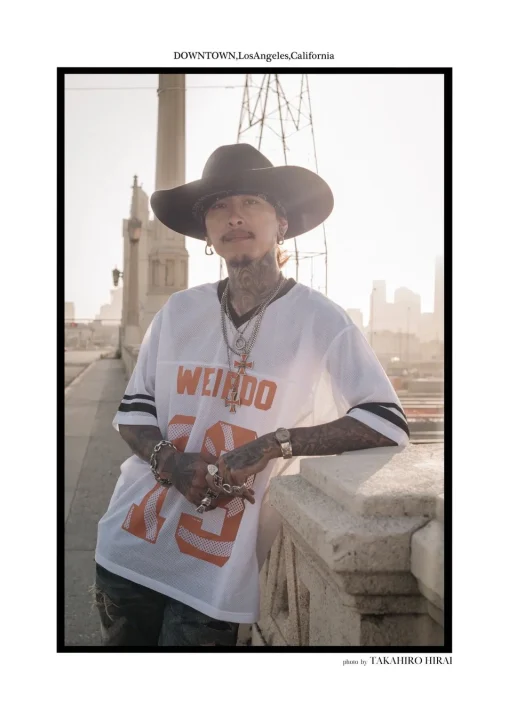 タトゥーのある男性がカウボーイハットとメッシュジャージを着用し、橋の欄干に寄りかかっています。背景にはロサンゼルスの街並みが広がっています。