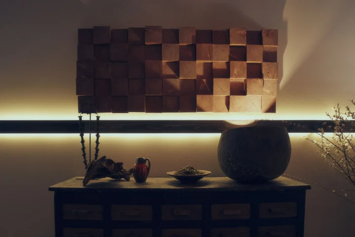 間接照明で照らされた木製ブロックの壁飾りの下に、コンソールテーブルが置かれ、花瓶や乾燥した葉の入った器、白い花の枝などの装飾品が並んでいます。落ち着いた暖かみのある和モダンなインテリア空間を捉えた一枚です。