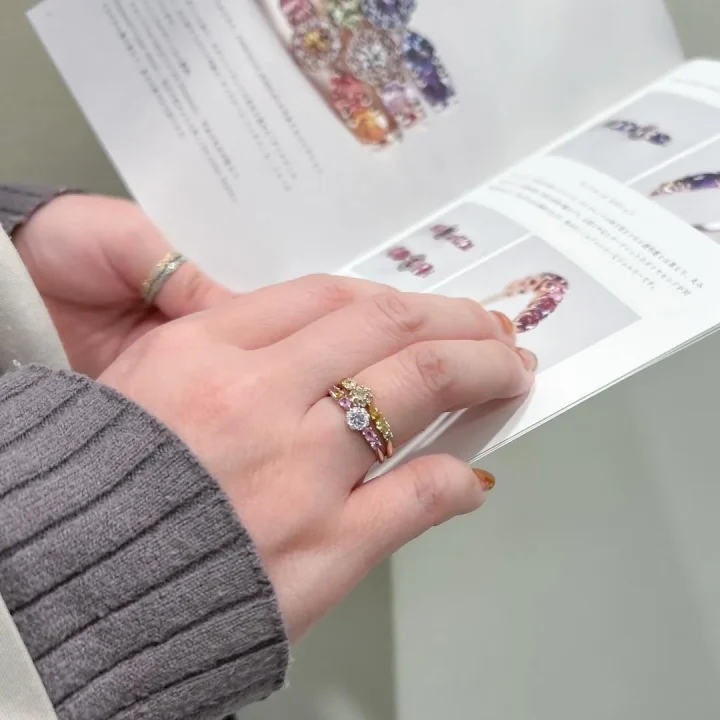 カラフルな宝石の指輪を試着中の手元と、ジュエリーのパンフレットが写っています。