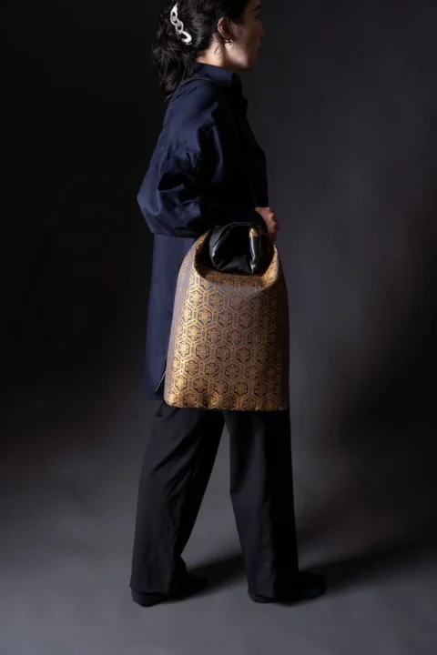 紺色のシャツと黒いパンツを着用した女性が、金色と黒の幾何学模様が特徴的なバッグを手にしている全身像です。