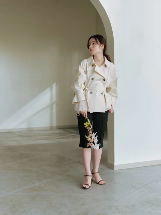 ベージュのショートトレンチコートと黒のキュロットを着用した女性が、屋内のアーチ状の空間で蘭の花束を持ち、左を向いて立っています。