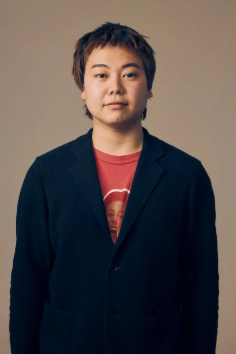 明るい背景を背に、黒いブレザーと赤いTシャツを着た若者が正面を見ているポートレート。短めの茶髪で、Tシャツには顔のグラフィックが描かれている。