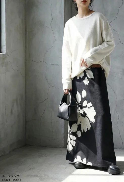 白いゆったりとしたセーターと、黒地に大きな白い花柄が特徴のロングスカートを着用した女性の全身コーディネート