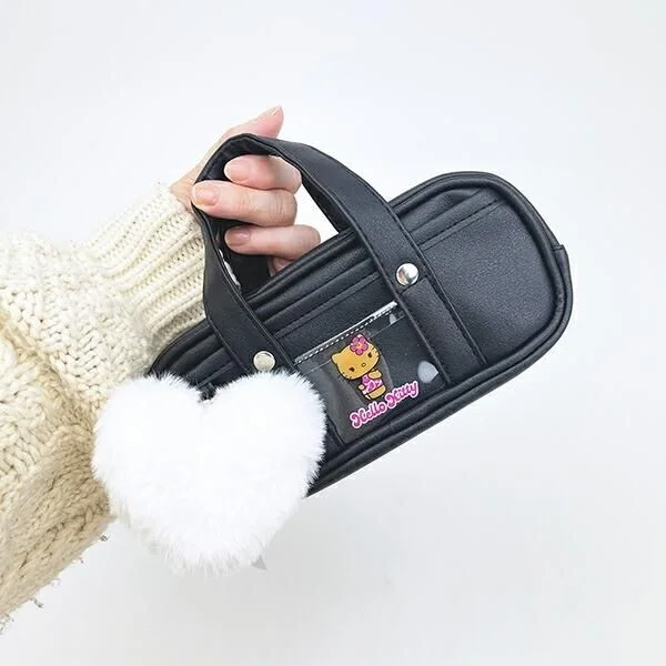 白いニットを着た手が、白いふわふわのハートチャームが付いた黒いハローキティデザインのポーチを持っている
