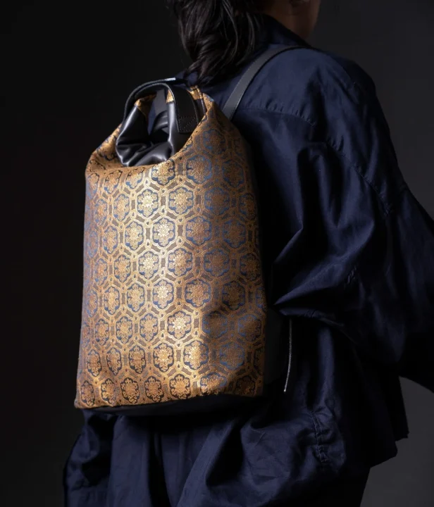 紺色の衣服を身につけた人物が、金色と紺色の伝統的な六角形模様のリュックサックを背負っている様子。