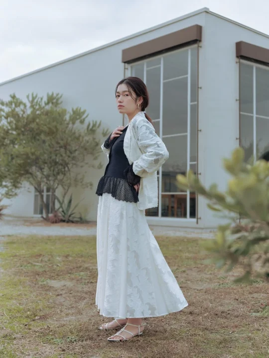 白いジャケットと黒いトップス、白いロングスカートを着用した女性が、屋外でモダンな白い建物を背景にポーズをとっています。