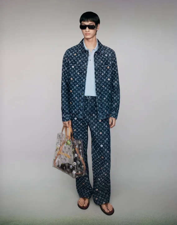 ルイ・ヴィトンのモノグラム柄デニムセットアップを着用した男性が、サングラスをかけ、クリアなトートバッグを持って立っている全身像です。