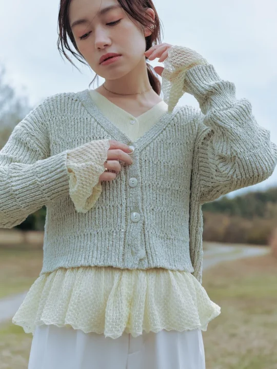 淡い色のカーディガンとフリルのトップスを着た若い女性が、自然の中のような場所でポーズをとっている。全体的に柔らかく、落ち着いた雰囲気のポートレート。