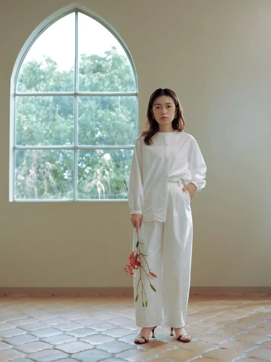 白いエンブロイダリーのブラウスとパンツのセットアップを着用した女性が、赤いユリの花を持ち、アーチ型の窓がある明るい室内で立つポートレート。