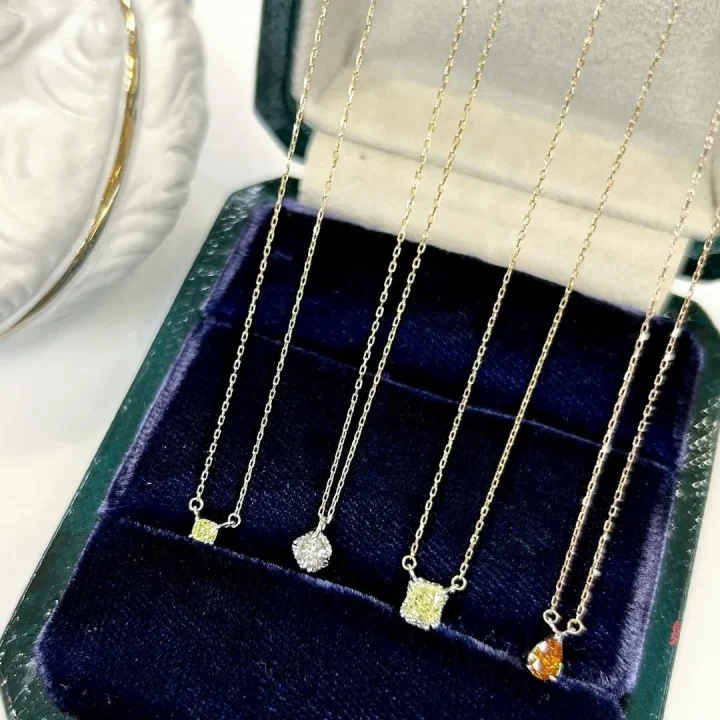 濃い青色のベルベットのジュエリーボックスに、イエロー、クリア、オレンジなど様々な色と形の宝石が付いた繊細なゴールドネックレスが4本並べられています。