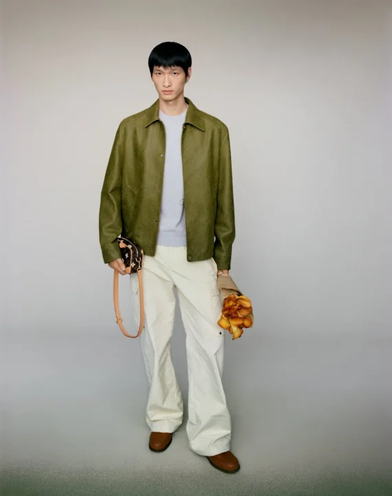 オリーブグリーンのジャケットとクリーム色のカーゴパンツを着用した男性モデルが、スタジオで全身を撮影されています。左手にはモノグラム柄のミニバッグ、右手にはオレンジ色の花束を持ち、カメラをまっすぐ見つめています。