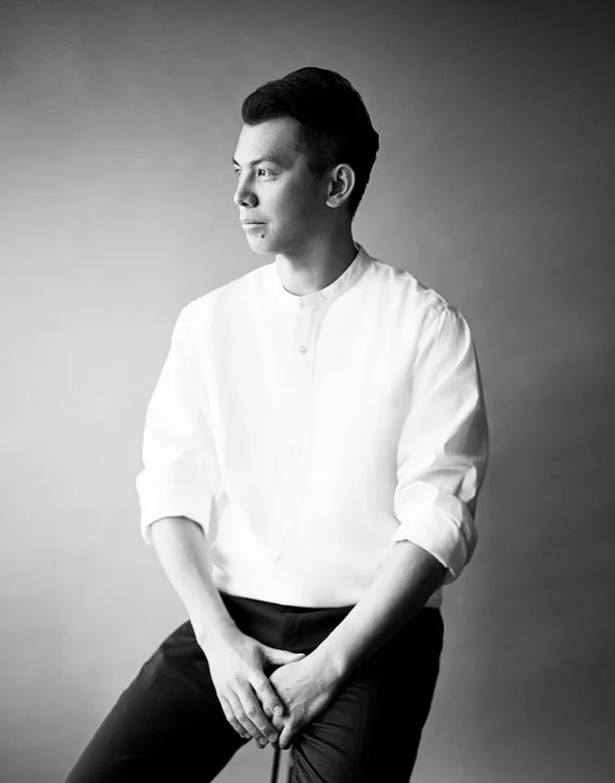 白いシャツを着た男性が椅子に座り、左方向を見つめているモノクロのポートレート写真です。両手を組み、落ち着いた表情をしています。