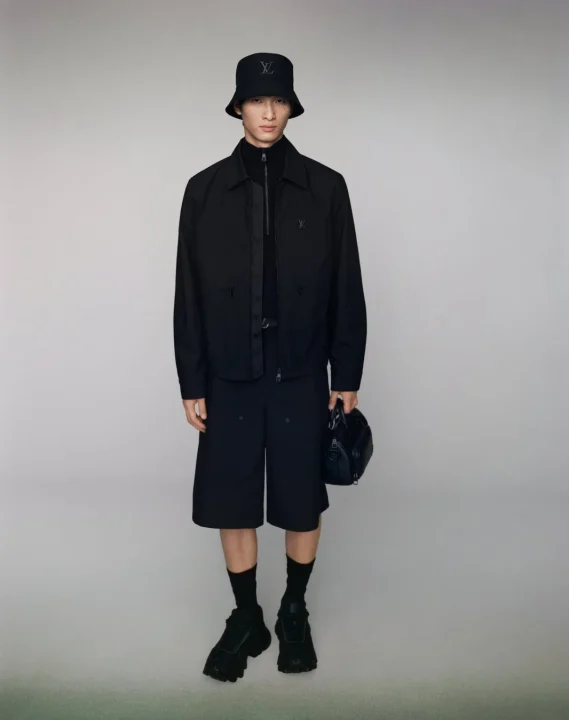 全身を黒で統一したルイ・ヴィトンのファッションを着用した男性モデルが写っています。バケットハット、ジャケット、ハーフパンツ、厚底スニーカーを身につけ、黒いハンドバッグを持っています。モダンでスタイリッシュな印象です。