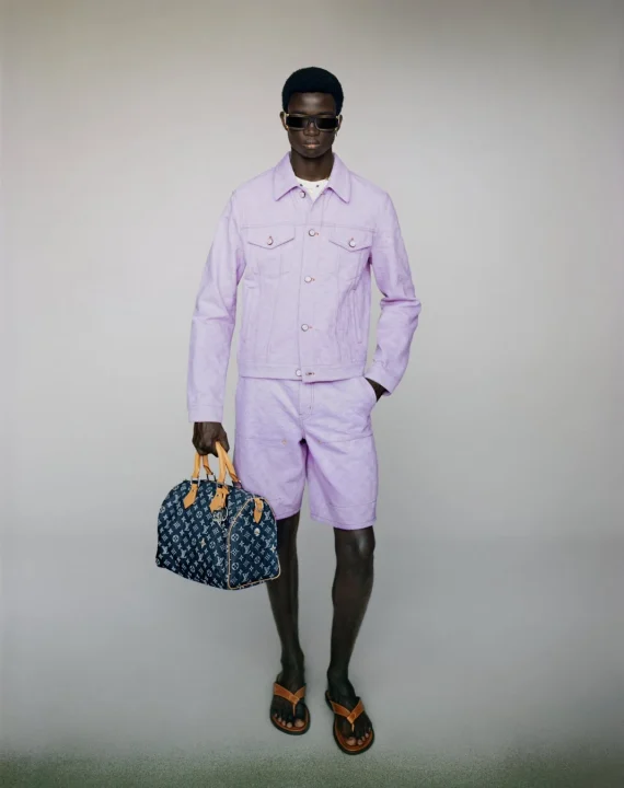 ライラック色のデニムジャケットとショートパンツを着用し、サングラスをかけた男性モデルが、ルイ・ヴィトンのバッグを持って立っている全身像。スタジオで撮影されたファッション写真です。