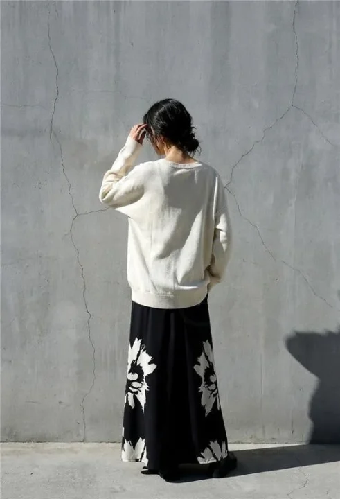 後ろ姿の女性が立っています。クリーム色のシンプルなセーターと、黒地に白の大きな花柄が特徴的なロングスカートを着用