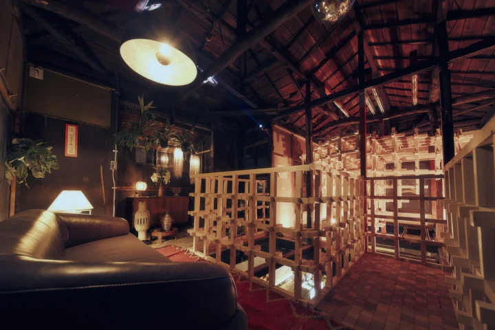 暗がりの室内空間で、革張りのソファ、観葉植物、大きな照明が配されています。中央には木材を組み合わせた独特のグリッド構造があり、天井にはミラーボールが吊るされています。どこか懐かしさを感じるレトロモダンな雰囲気が特徴です。