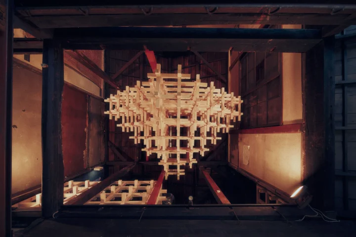 古い建築物のような空間に吊り下げられた、無数の木片が複雑に組み合わされた幾何学的な構造物です。温かい光に照らされ、独特の雰囲気を醸し出しています。