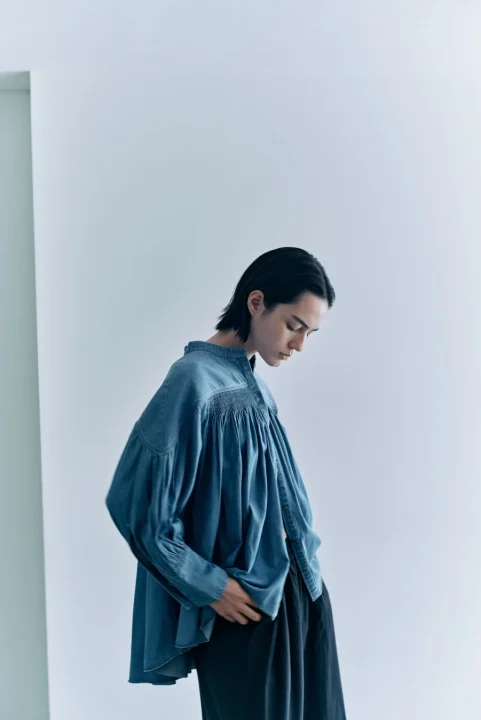 ゆったりとした青いスモッキングブラウスと黒いワイドパンツを着用した人物