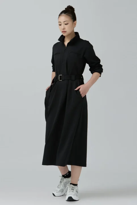 黒の襟付きミディ丈ワンピースにベルトを合わせ、白いスニーカーを履いた女性が、両手をポケットに入れてポーズをとっているスタジオ写真。カジュアルながらも洗練されたスタイルです。