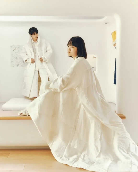 白を基調としたミニマルな空間で、キルティング素材のオーバーサイズな白い衣装を着用した2人のモデルがポーズをとるファッション写真。清潔感とモダンな雰囲気が特徴です。