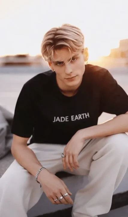 金髪の若い男性が屋外で座り、カメラを見つめています。黒いTシャツと白いパンツを着用し、アクセサリーを身につけています。夕焼けの光が背景に広がる、スタイリッシュなポートレートです。