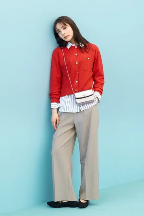 ライトブルーの壁にもたれかかる女性の全身ショット。赤のカーディガンにストライプシャツ、ベージュのワイドパンツを合わせた、春らしいきれいめカジュアルなコーディネートです。