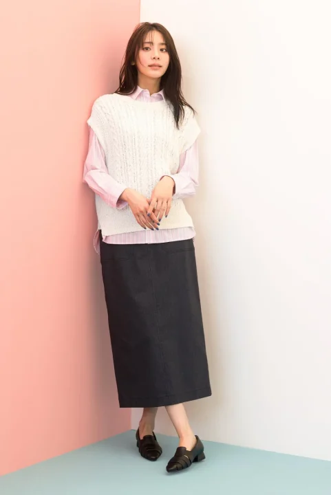 ピンクと白の壁を背景に立つ女性の全身写真です。白いニットベスト、ピンクのストライプシャツ、黒いロングスカート、黒い靴を着用し、カメラを見つめています。