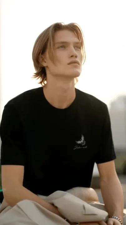 黒いTシャツを着たロングヘアの若い男性が、屋外で空を見上げているポートレート。自然光が当たり、穏やかな雰囲気の画像です。