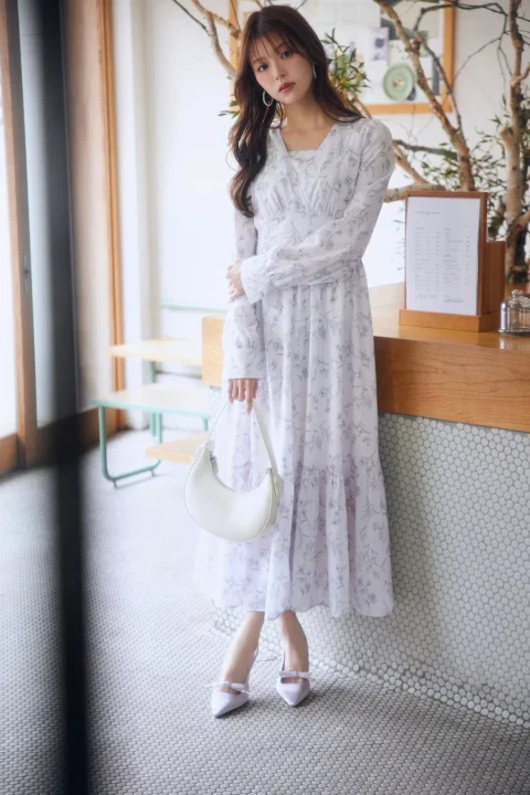 明るいカフェのような空間で、花柄の白いロングワンピースを着た女性が立っています。白いバッグと淡いパープルのパンプスを合わせた、上品でフェミニンな雰囲気のコーディネートです。