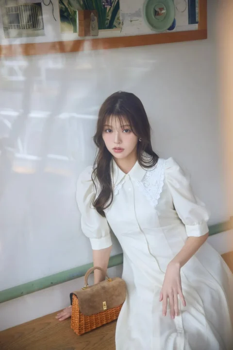 白いパフスリーブのワンピースを着用した若い女性が、ブラウンのスエードとラタン素材のバッグを傍らに置いて座っているポートレート。上品で落ち着いた雰囲気のファッション写真です。