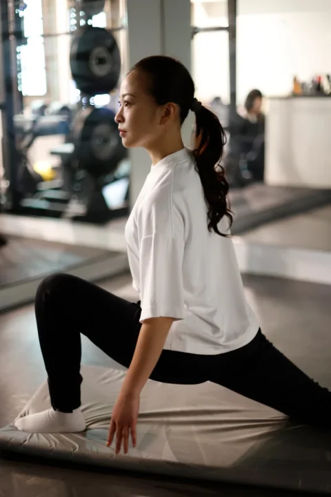ジムのような場所で、白いTシャツと黒いレギンスを着用した女性が、マットの上でストレッチをしている様子。健康的なライフスタイルを示すシーンです。