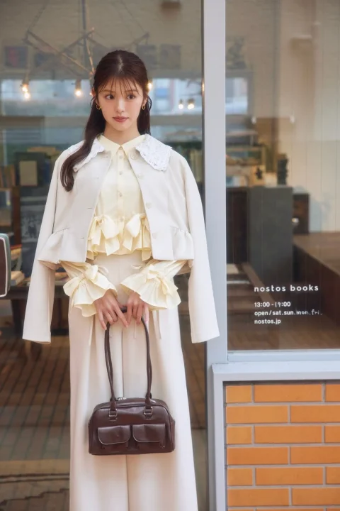 フリルとレースがあしらわれたベージュのトップスと白いアウターを着用した女性が、茶色のハンドバッグを持ち、書店「nostos books」の前に立っている様子を捉えた画像です。