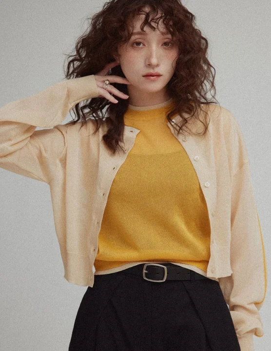 黄色のトップスとベージュのカーディガン、黒いパンツを着用し、指にリングをはめてポーズをとる女性